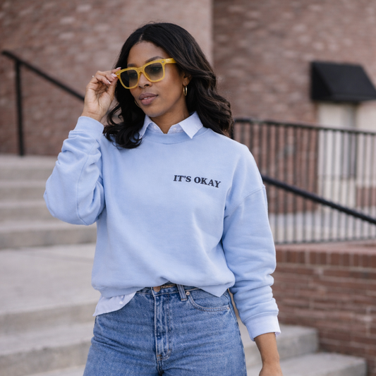 Woman wearing light blue It’s Okay embroidered sweatshirt with jeans and tote bag styled in casual streetwear look for Shop Jayla.